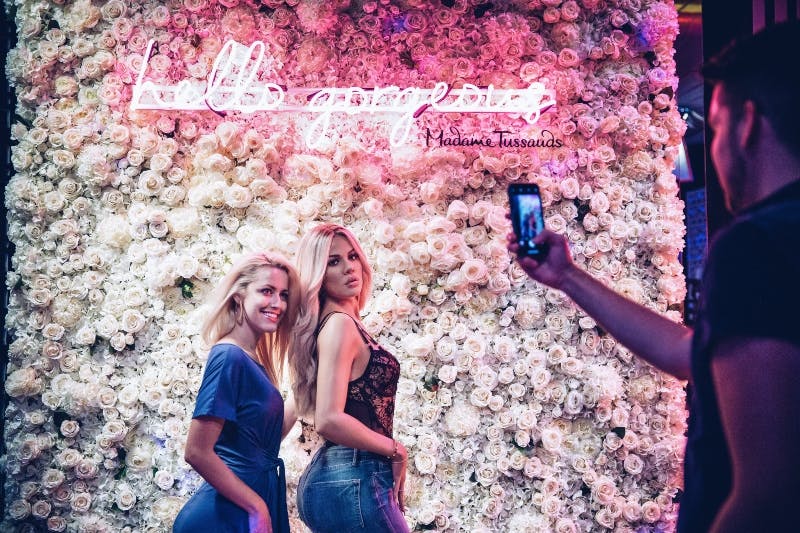 화려한 인사말과 마담 투소 문구가 적힌 네온 사인이 있는 꽃무늬 벽 앞에서 두 사람이 포즈를 취하고 있습니다.