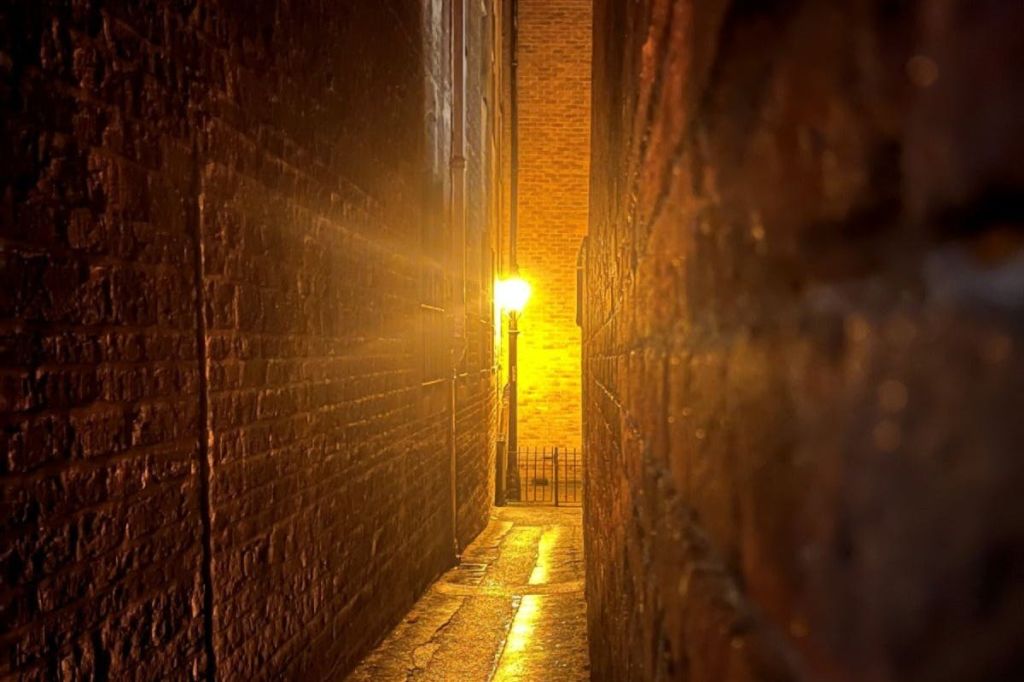 따뜻한 가로등이 비추는 좁은 골목길로, 양쪽에 젖은 반사 표면과 벽돌 벽이 있습니다.