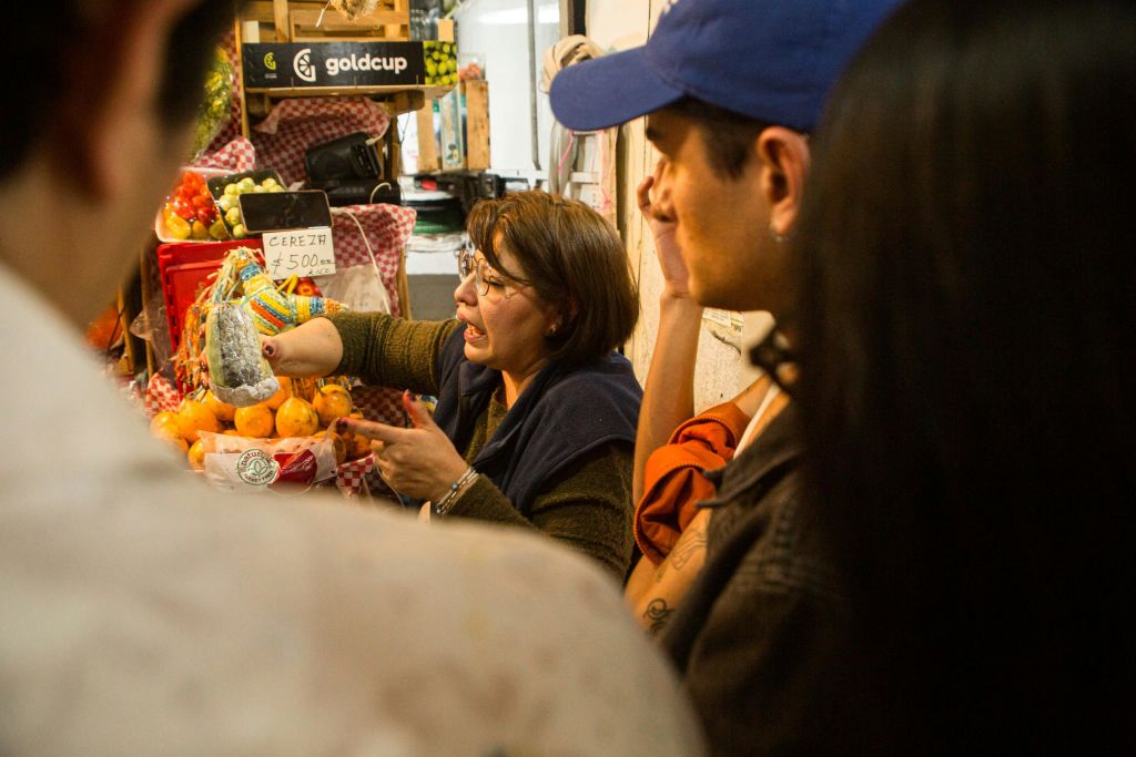 시장 가판대에 있는 여성은 고객과 상호 작용하며 과일을 진열하고 안경을 착용하는 반면, 다른 여성은 아이템을 관찰하거나 선택합니다.