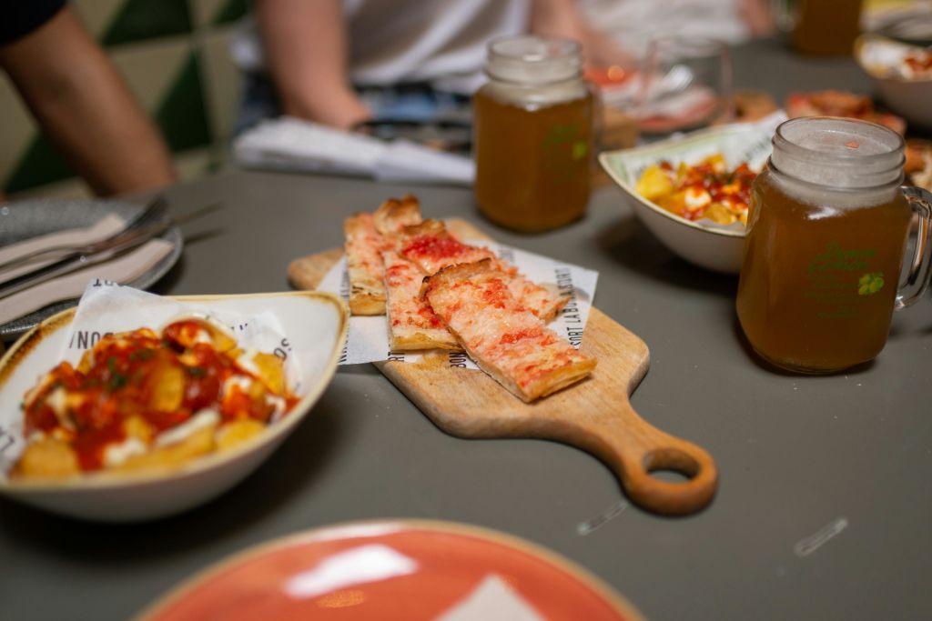 회색 테이블 위에 음식과 음료를 놓은 나무 판에 피자 조각이 놓여 있습니다. 사람들이 배경에 앉아 있습니다.