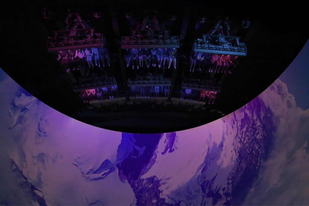 보라색과 흰색 산악 지형이 표시된 커다란 곡선형 화면 위에 서스펜션된 차량 서비스를 이용하는 사람들.