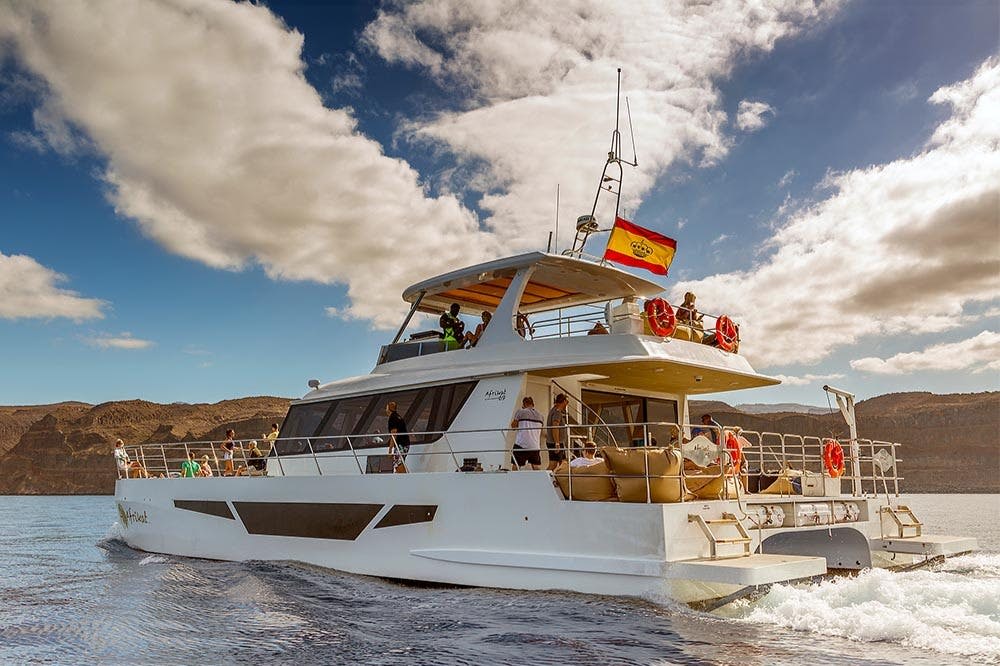 사람들이 탑승한 커다란 흰색 요트가 물 위를 항해하고 있습니다. 요트는 부분적으로 흐린 하늘 아래 빨간색과 노란색 깃발을 표시합니다.