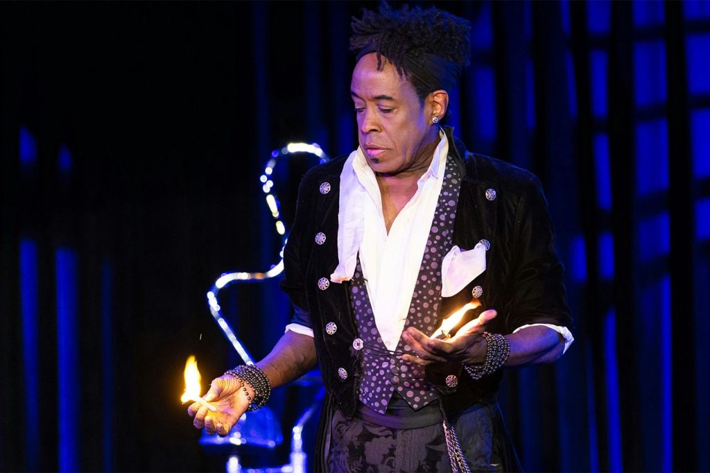 파란색 조명이 있는 어두운 무대에서 양손에 불을 들고 있는 정교한 복장을 한 연주자.