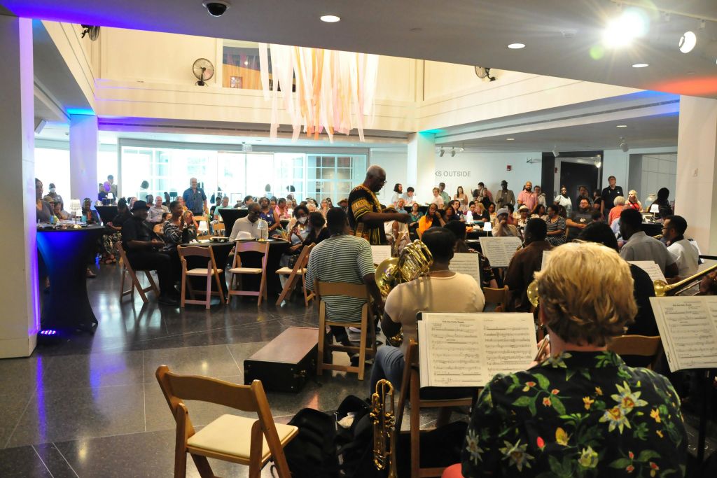 원형 테이블에 앉은 관객이 있는 넓고 조명이 밝은 방에서 밴드가 공연하고, 지휘자가 음악가들을 이끌고 있습니다.