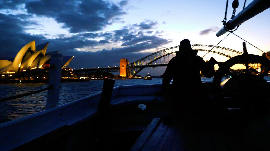 도시의 불빛과 흐린 하늘을 배경으로 해질 무렵 시드니 하버 브리지 앞에서 보트를 탄 사람의 실루엣.