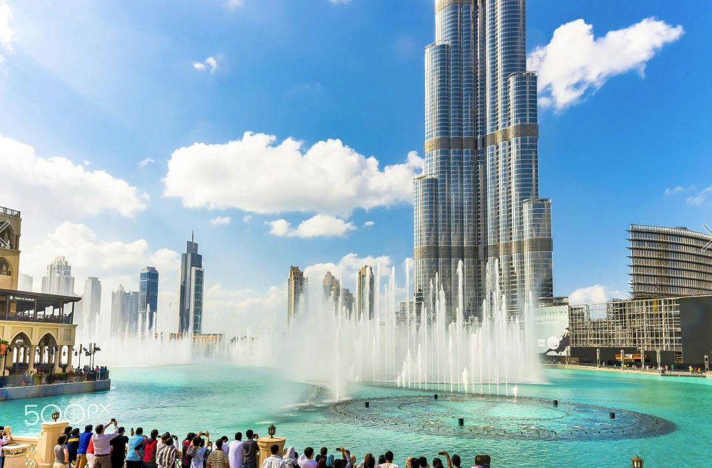 맑고 화창한 날 하늘에 구름이 몇 개 있는 높은 고층빌딩 앞에서 대형 분수쇼를 관람하는 관광객들.