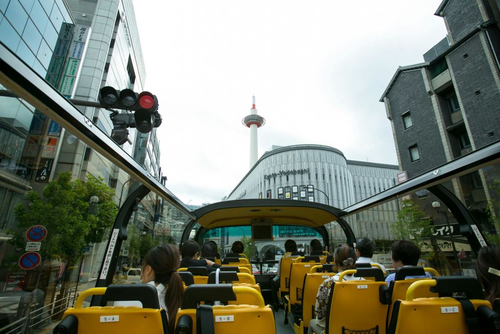 현대적인 건물과 높은 전망대를 배경으로 한 도시의 오픈탑 투어 버스에 앉아 있는 사람들.