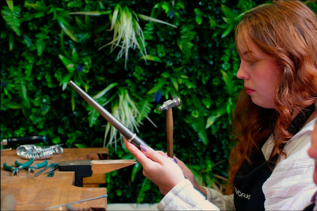 길고 붉은 머리카락을 가진 사람은 울창한 녹색 식물을 배경으로 망치와 함께 금속 도구를 사용합니다.