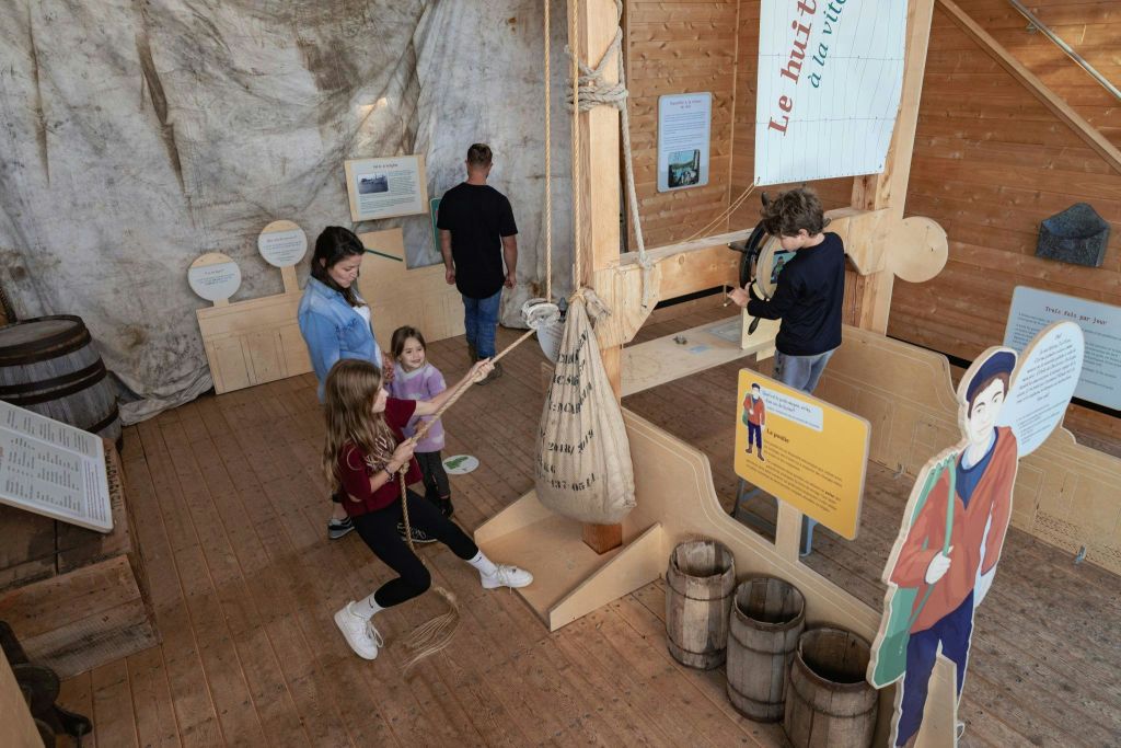 목조 건물 안에 있는 어린이와 성인. 두 명의 소녀는 자루에 부착된 밧줄을 당기고, 두 명의 성인은 디스플레이를 관찰합니다.