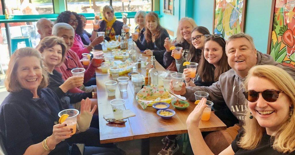 밝은 색상의 실내 환경에서 긴 테이블에 둘러앉아 미소를 짓고 음료를 들고 있는 사람들의 모임.