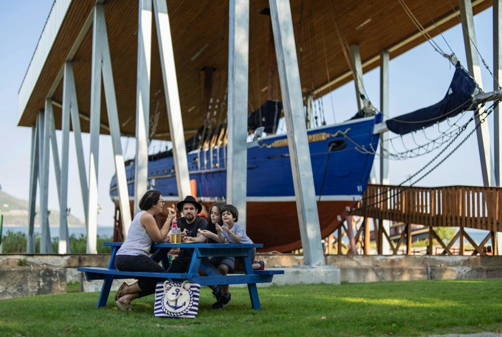 4인 가족이 기둥에 진열된 커다란 나무 배 아래 파란색 피크닉 테이블에 앉아 있습니다. 바닥에 줄무늬 가방이 있습니다.