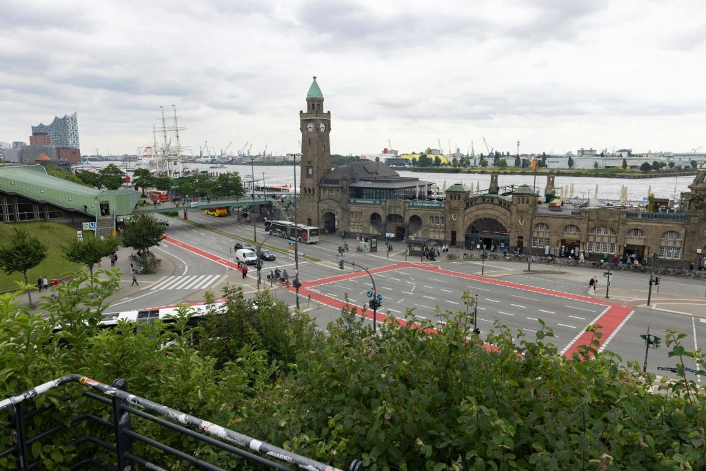 분주한 교차로 옆에 있는 시계탑과 벽돌 건물에서 흐린 하늘 아래 크레인과 배가 있는 항구가 내려다보입니다.