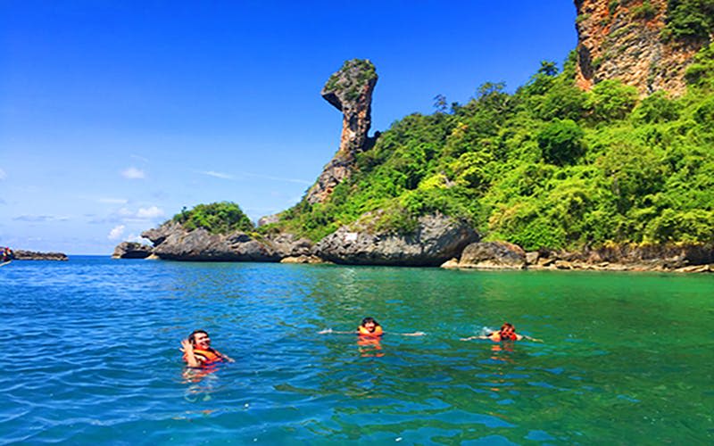세 사람은 높은 바위 근처의 맑고 푸른 물에서 수영하고 밝고 푸른 하늘 아래 무성한 녹색 절벽에서 수영합니다.