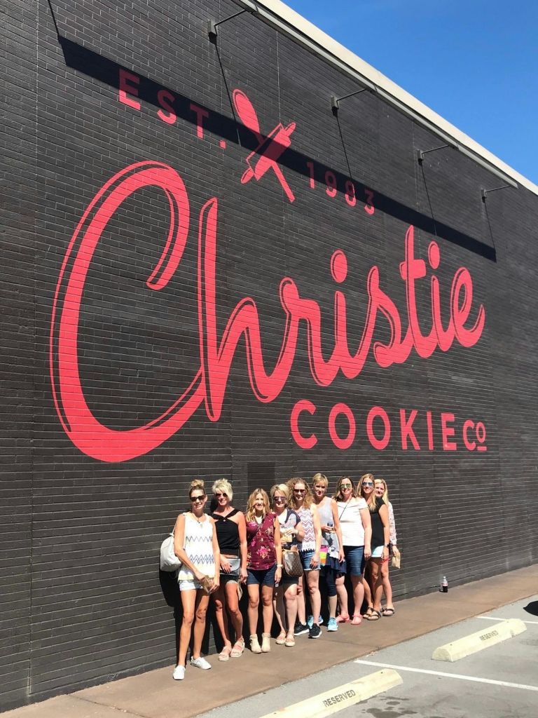 빨간 글씨로 'Christie Cookie Co.' 라는 글자가 적힌 벽돌 벽 앞에서 포즈를 취하고 있는 한 무리의 사람들.