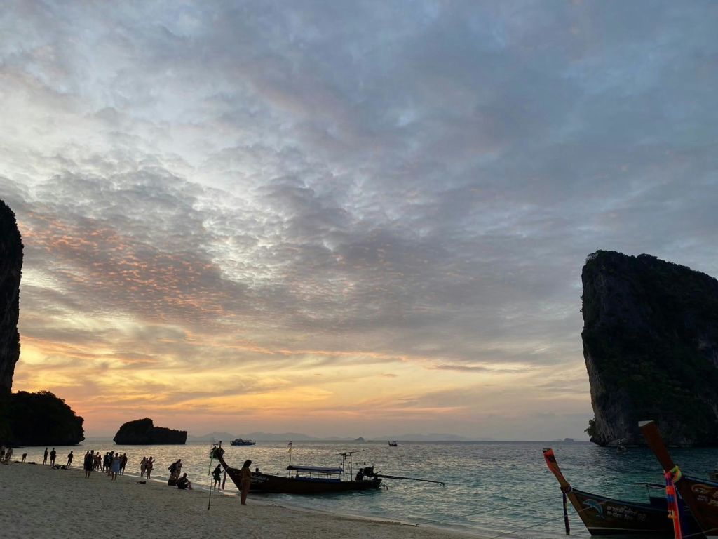 해안의 보트와 커다란 암석을 배경으로 석양을 배경으로 해변을 거닐고 있는 사람들.
