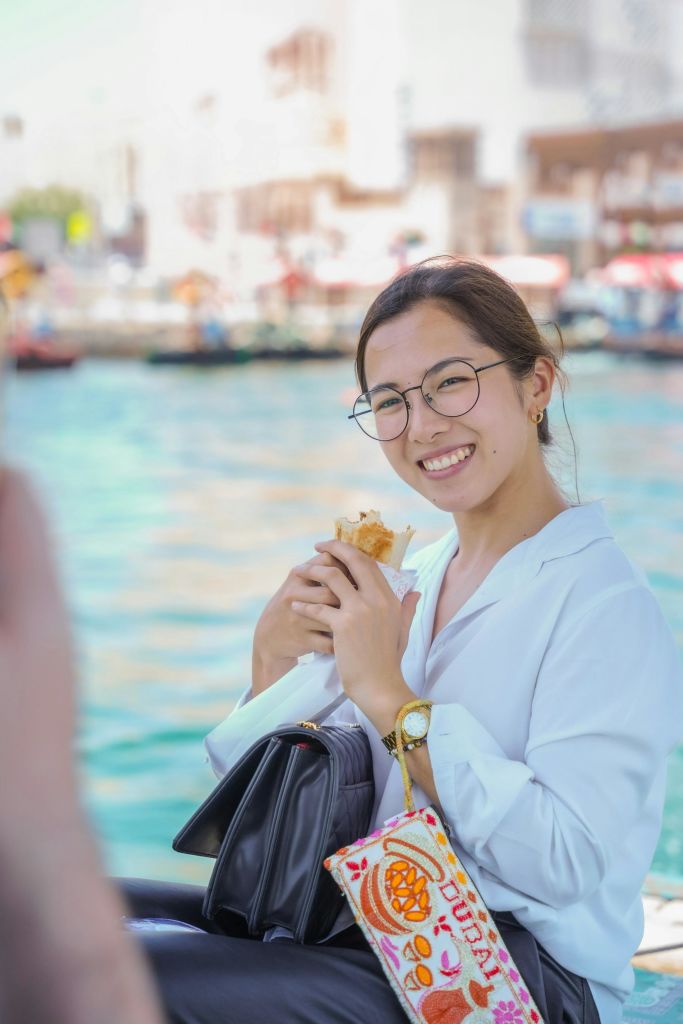 안경을 쓰고 샌드위치와 카메라를 들고 웃고 있는 한 여성이 보트와 건물을 배경으로 바닷가 근처에 서 있습니다.