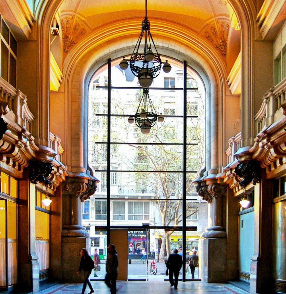 아치, 커다란 창문, 샹들리에, 출구를 향해 걸어가는 사람들이 있는 화려한 실내 복도. 야외에 도시 거리가 보입니다.