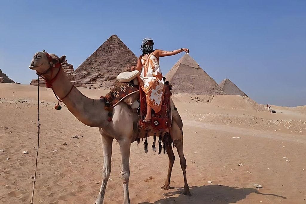 한 사람이 맑고 푸른 하늘 아래 사막 풍경의 피라미드 근처에서 낙타를 타고 있습니다.