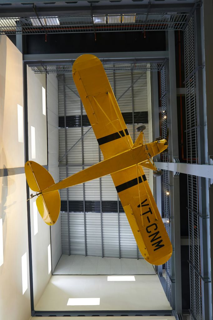 검은 줄무늬와 등록 VT-CNM이 있는 노란색 소형 항공기가 현대적인 실내 공간의 천장에 매달려 있습니다.