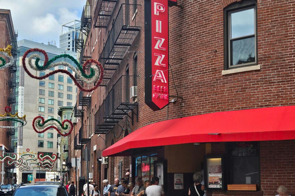 도시 거리의 입구 근처에 수직 피자 간판, 붉은 천막, 사람들이 모여 있는 붉은 벽돌 건물.