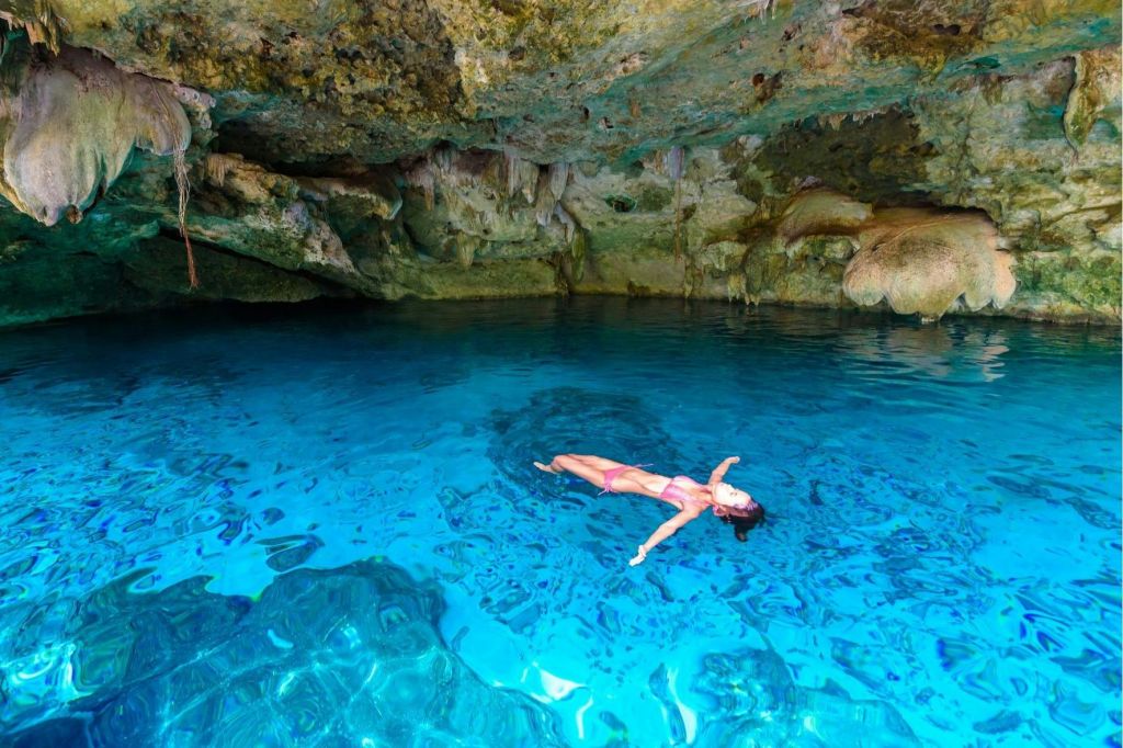 한 사람이 바위 벽과 종유석이 있는 동굴 안의 맑고 푸른 물에 등을 대고 떠 있습니다.