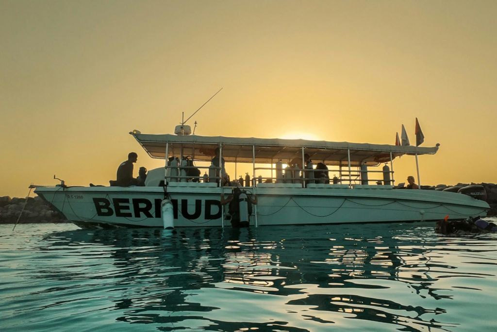 일몰에 잔잔한 물 위에 '버뮤다' 라는 라벨이 붙은 보트에는 선상에서 볼 수 있는 사람들의 실루엣이 있습니다.