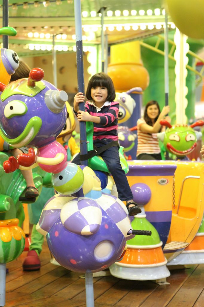 줄무늬 셔츠를 입은 어린이가 알록달록한 곤충 테마의 회전목마를 타고, 또 다른 어린이와 어른이 웃고 있습니다.