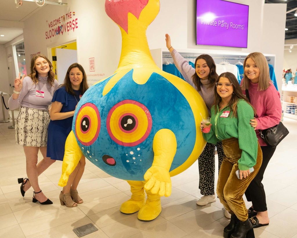 네 명의 여성이 실내 행사에서 크고 화려한 라운드 마스코트와 함께 포즈를 취하고 있으며, 배경에는 "Private Party Rooms!" 사인이 있습니다.