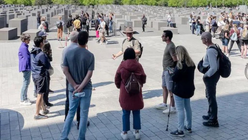 한 무리의 사람들이 수많은 콘크리트 블록이 있는 기념관 근처에서 투어 가이드의 말을 듣고 있습니다. 배경에는 더 많은 방문자가 보입니다.
