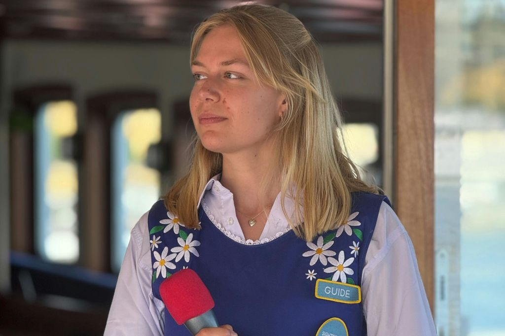 꽃무늬 디테일이 있는 파란색 조끼를 입고 빨간색 마이크와 '가이드' 라는 이름표를 들고 있는 금발의 여성.
