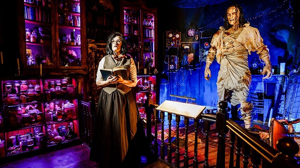 으스스한 실험실에서 책을 들고 있는 빅토리아 시대의 옷을 입은 여성. 백그라운드에서 만나는 프랑켄슈타인의 실물 크기 괴물