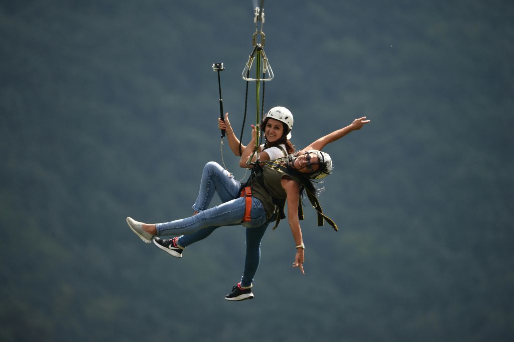 헬멧, 하네스, 캐주얼 복장을 모두 착용하고 한 명은 셀카봉을 들고 숲 위에 집라인을 켠 두 사람.