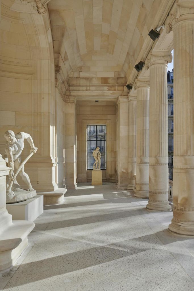 도구로 구부러진 남자와 멀리 서 있는 형상을 포함한 고전적인 조각상이 있는 햇살이 비추는 콜로네이드.