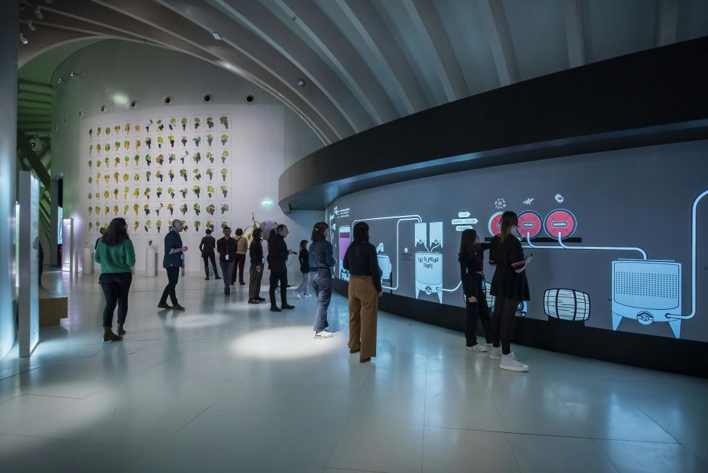 흰색 벽과 예술적인 설치물이 있는 현대적이고 넓은 객실에서 사람들은 커다란 곡선 디지털 디스플레이와 상호 작용합니다.