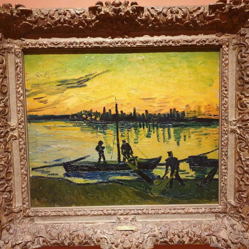 노란색과 초록색 하늘 아래 저 먼 도시의 스카이라인을 배경으로 석양이 질 무렵 강가에서 배를 내리는 화려한 액자의 그림.