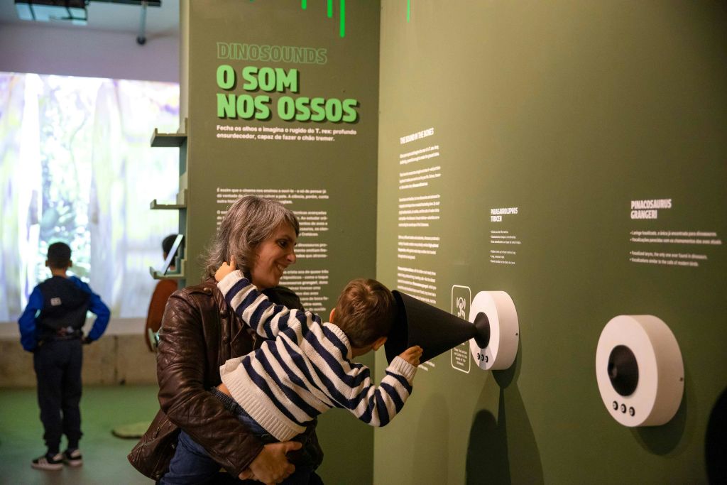 한 여성이 "Dinossounds, O Som nos Ossos" 라고 표시된 박물관의 벽에 부착된 콘을 통해 듣고 있는 아이를 안고 있습니다.