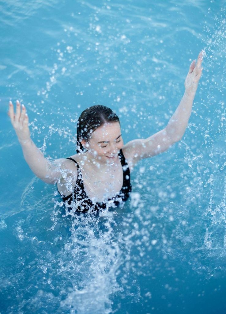 검은색 수영복을 입은 사람이 수영장에 서서 팔을 들어 기쁘게 물을 튀기고 있습니다.