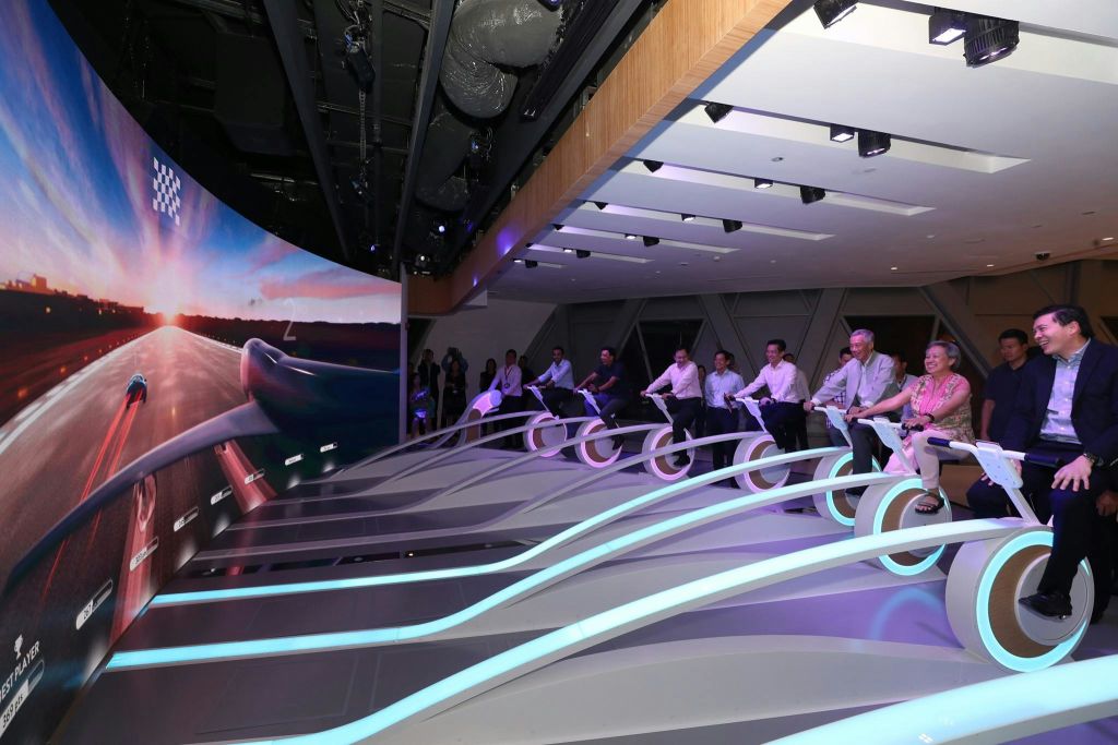 비즈니스 복장을 한 사람들은 실내에서 조명이 켜진 바퀴가 달린 미래형 고정식 자전거를 타고 소규모 청중의 시선을 받습니다.