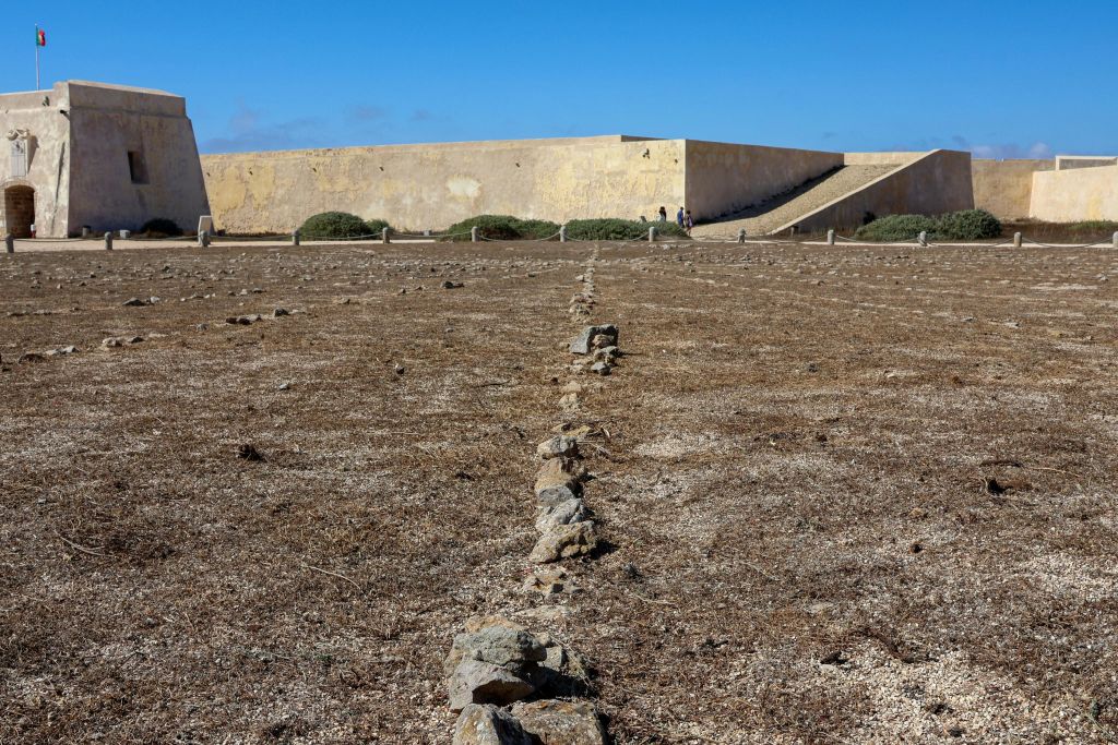 맑고 푸른 하늘 아래 돌로 된 요새 벽으로 이어지는 돌들이 줄지어 있는 메마르고 척박한 땅.
