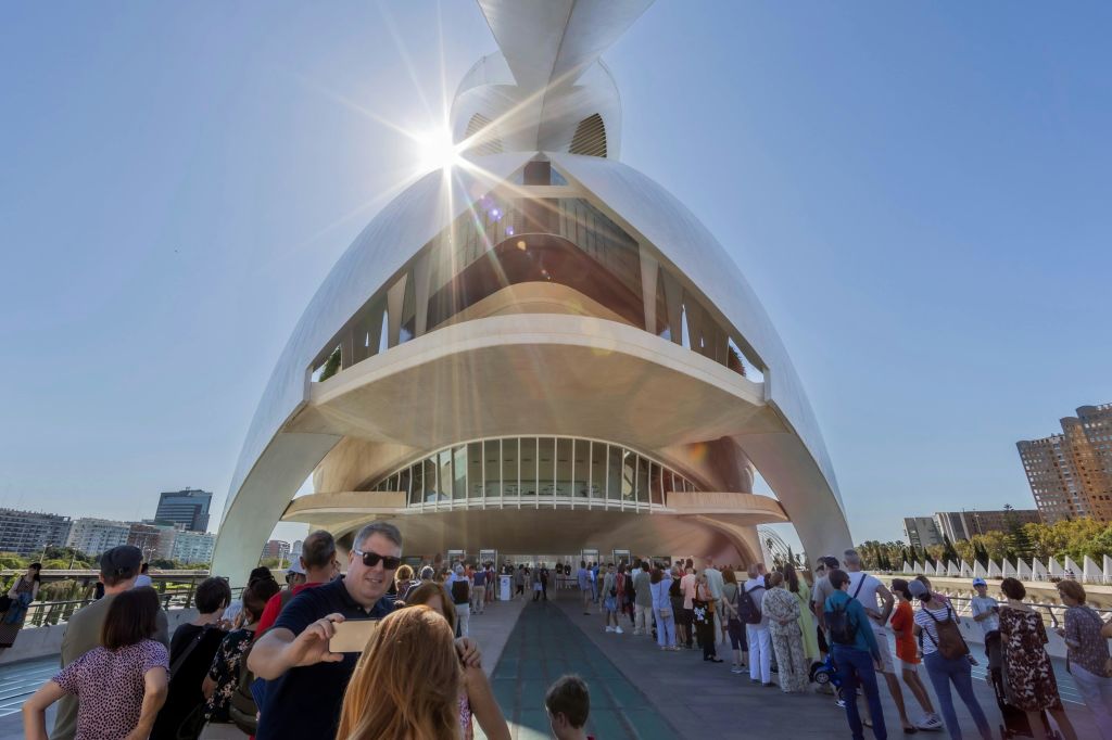 맑고 화창한 푸른 하늘 아래 곡선형 디자인의 미래지향적이고 하얀 건물 밖에서 한 무리의 사람들이 줄을 서서 기다립니다.