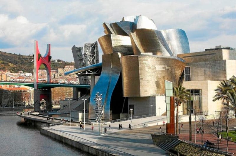 강변 산책로 옆에 곡선형의 반사 표면이 있는 현대적인 금속 건물. 사람들은 아래 길을 걷습니다.