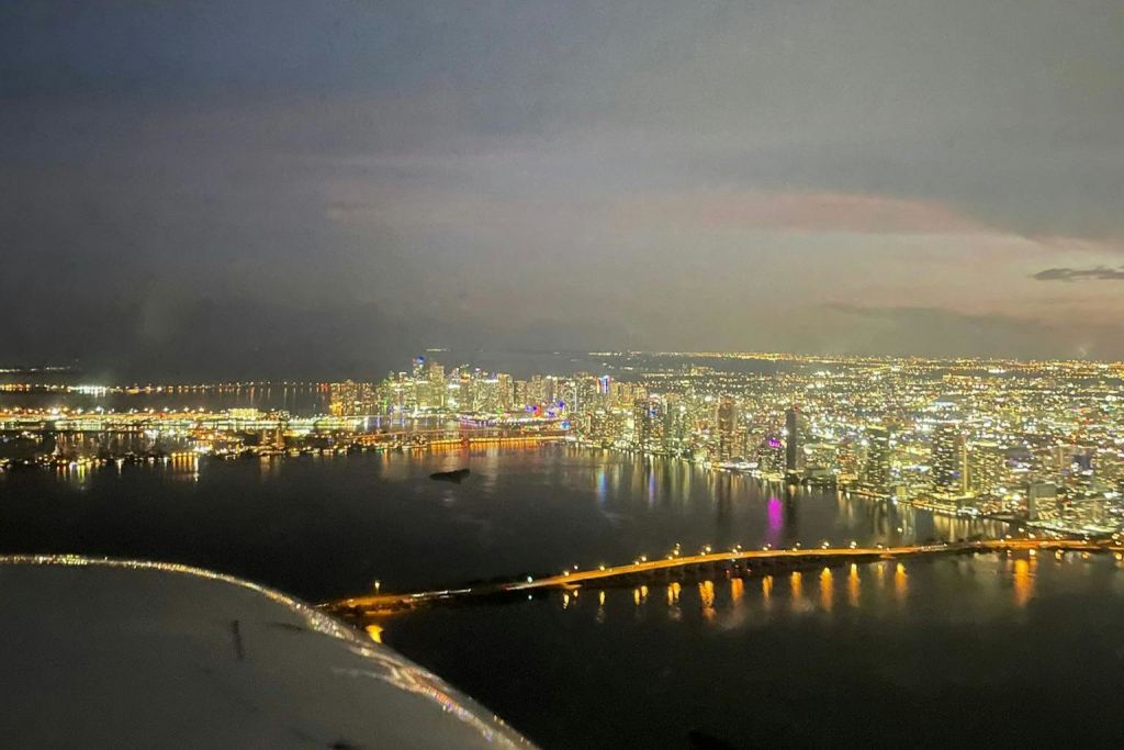 밤에는 조명이 켜진 건물, 물 위에 놓인 다리, 수면에 반사되는 조명이 있는 도시 스카이라인의 공중 전망을 감상할 수 있습니다.