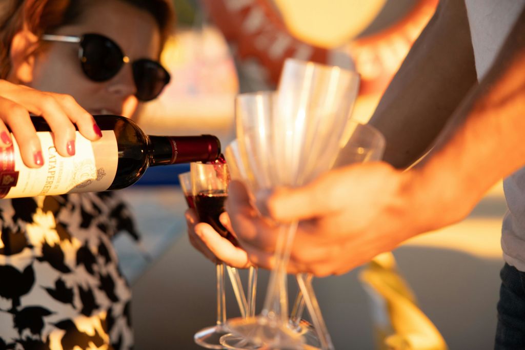 선글라스를 쓴 사람이 병에 담긴 레드 와인을 다른 사람이 들고 있는 여러 개의 투명한 잔에 붓고 있습니다.