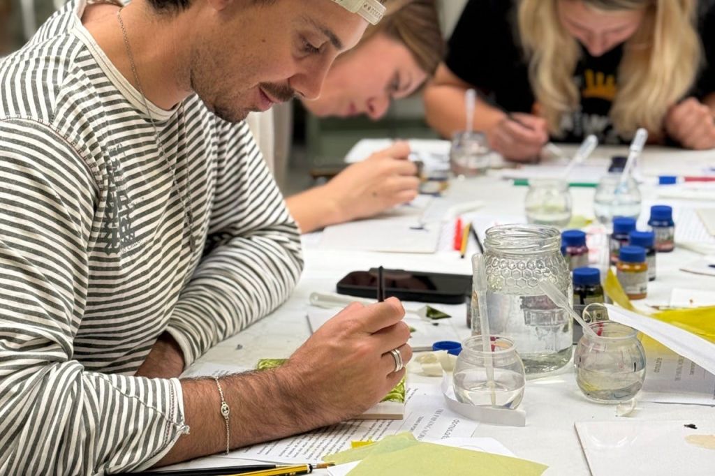 다양한 재료와 도구를 테이블에 올려놓고 세부적인 아트워크나 글쓰기 작업에 중점을 두고 테이블에 앉아 있는 사람들.