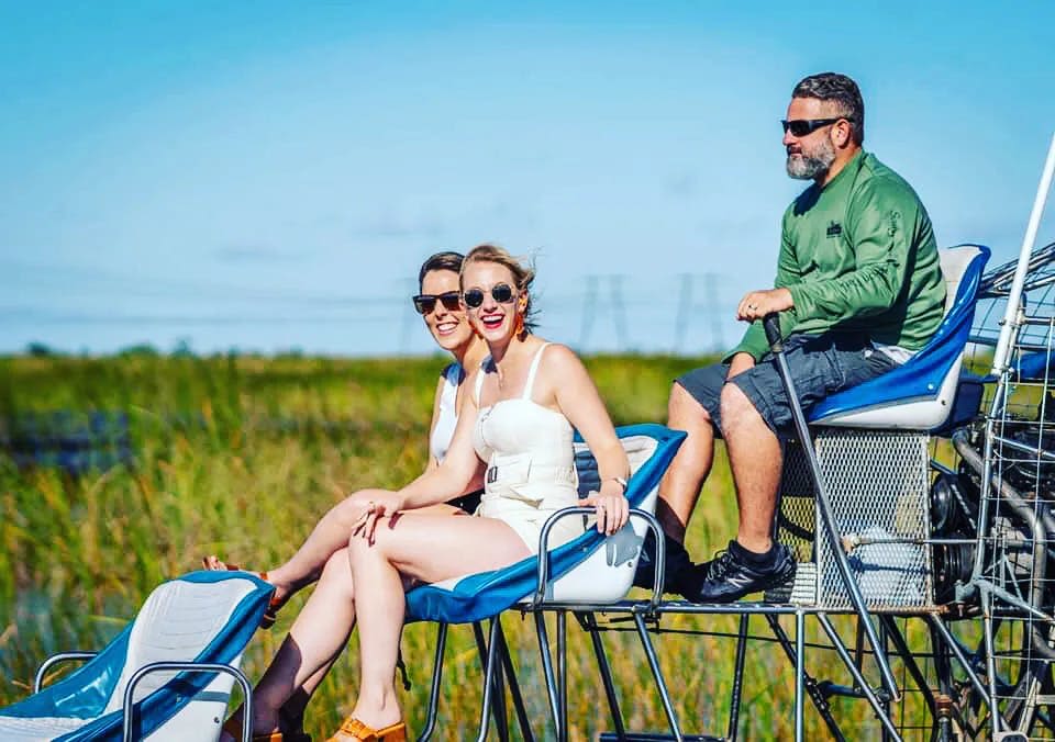 두 여성과 한 남성이 에어보트를 타고 맑고 푸른 하늘 아래 잔디가 우거진 습지대를 지나고 있습니다.