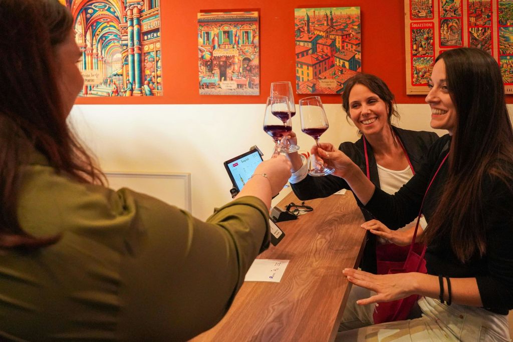 나무 조리대 뒤에서 사람과 함께 레드 와인을 마시며 웃고 있는 두 여성. 주황색 벽에 화려한 그림.