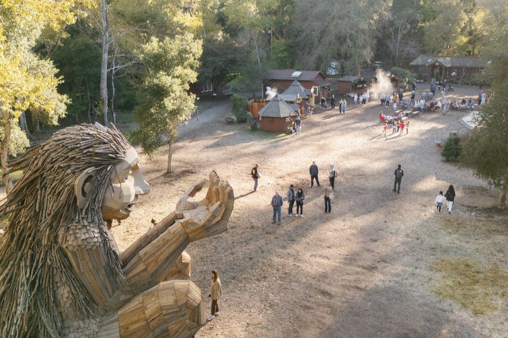커다란 나무 조각상, 근처를 걷는 사람들, 숲이 우거진 작은 건물이 있는 야외 풍경.