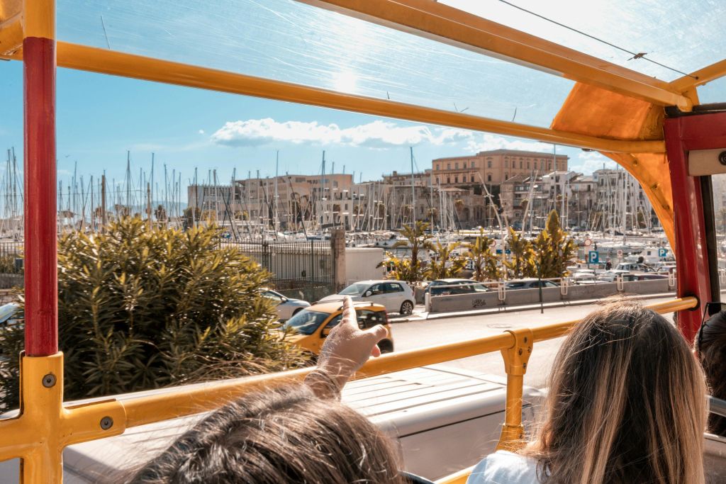 투어 버스의 상부 갑판에 앉아 보트와 건물을 배경으로 항구를 가리키는 사람들.