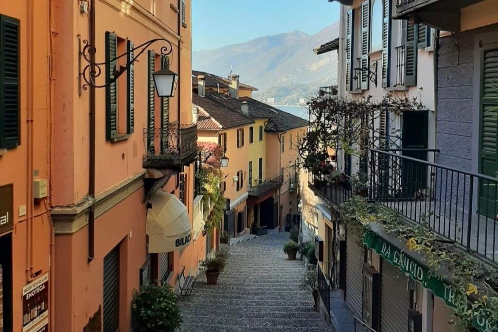 셔터, 식물, 등불이 있는 화려한 건물 사이로 좁은 자갈길이 있고, 맑은 하늘 아래 산이 한눈에 들어옵니다.