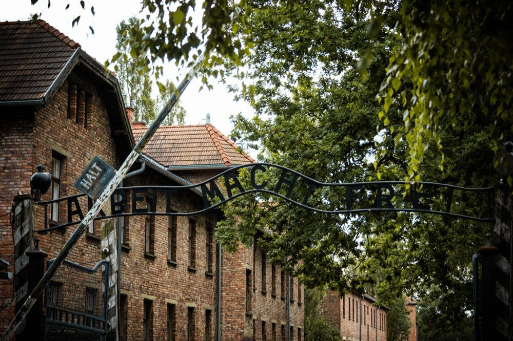 붉은 벽돌 건물과 푸른 나무로 둘러싸인 길 위에 "Arbeit Macht Frei" 라고 새겨진 철문이 있습니다.